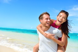 Happy couple in love on beach summer vacations. Joyful Asian girl piggybacking on young Caucasian boyfriend playing and having fun in sunny tropical destination for travel holiday.