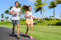 Sport couple exercising running outside on street in summer. Happy active young fit adults jogging together with tropical background in city park or resort road. Asian and Caucasian people.