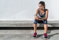 Fitness runner on mobile smart phone app tracking progress listening to music with earphones for fitness motivation. Athlete runner in sportswear relaxing sitting getting inspired. Asian mixed race.