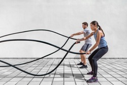 Fitness people exercising with battle ropes at gym. Woman and man couple training together doing battling rope workout working out arms and cardio for cross fit exercises.