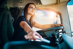 Portrait of beautiful young woman professional truck driver sitting and driving big truck. She is dangerously trying to take smart phone while driving.