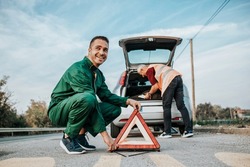 Two road assistant workers in towing service trying to fix car engine. One worker in foreground placing safety triangle on the road while another one doing something in background. 