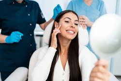 Beautiful and happy young woman sitting in medical chair and looking in the mirror. She is satisfied after successful beauty treatment with hyaluronic acid fillers or botulinum toxin injections.