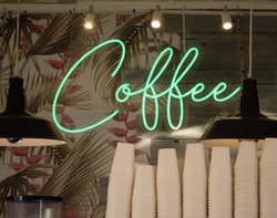 Cursive green neon sign COFFEE in front of palm and flower wallpaper and wood boards behind stacked paper cups and lamps