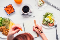 Morning habits of successful people. Day planning and healthy meal. Woman eating carrot and writing in notebook on the served for breakfast white wooden table. Top view, Selective focus
