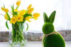 Cute green grass bunny rabbit and bouquet of fresh spring yellow tulips and daffodils flowers on the white kitchen table near the window. Happy Easter festive card. Selective focus. Copy space.
