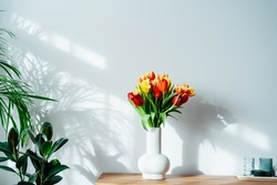 Scandinavian home interior with spring colorful bouquet of tulip flowers in ceramic vase standing on a wooden cabinet. Minimalist design with green plants and white wall. Biophilia style. Springtime.