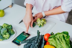 Go vegan. Close up woman cutting broccoli while cooking healthy dish according to the active online mobile application with Vegan diet program on her phone. Healthy lifestyle, weight loss concept