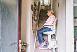 Senior woman using automatic stair lift on a staircase at her home. Medical Stairlift for disabled people and elderly people in the home. Selective focus.