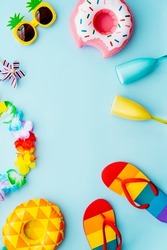 Flat lay of summer vibes concept with colorful pool party items, funny sunglasses, cocktail glasses, pineapple and donut inflatable drink holders, flip flops and flower necklaces on blue background