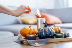 Female hand lighting a candle with a match in autumn cozy composition for hygge home mood. Pumpkins and candles on the tray with gray napkin on the coffee table in the living room. Selective focus.