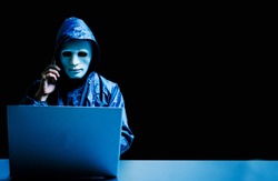Anonymous computer hacker in white mask and hoodie. Obscured dark face using laptop computer for cyber attack and calling on cellphone, Data thief, internet attack, darknet and cyber security concept.