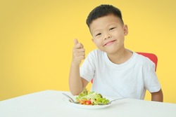 Asian 5 years old kindergarten boy child eating salad show thumb up, Kid enjoying Vegetarian Food with copy Space, Healthy food, healthy life style isolated on yellow background with clipping path