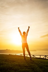 Successful sporty woman raising arms towards golden beautiful sunset and sea. Female athlete celebrating sport success and goals. Healthy lifestyle concept.