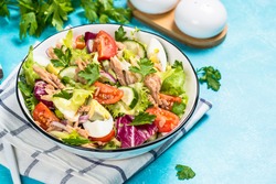Tuna salad with green leaves, eggs and vegetables in white bowl at blue background.