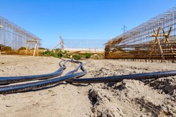 Long black plastic water pipes with blue stripe are placed on ground at building site in the background is bridge skeleton under construction.