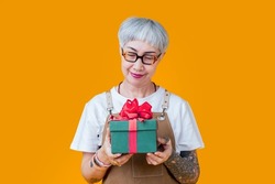 old senior asian female woman with glasses and tattoo wear brown apron hand holding present gift box to camera,smiling cheerful elder woman prepare present green box red ribbon isolate on yellow color
