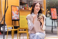 attractive asian woman pleased positive casual hand hold smartphone enjoy leisure relas sitting in front of street food booth corner in outlet department store mall relax shopping lifestyle concept