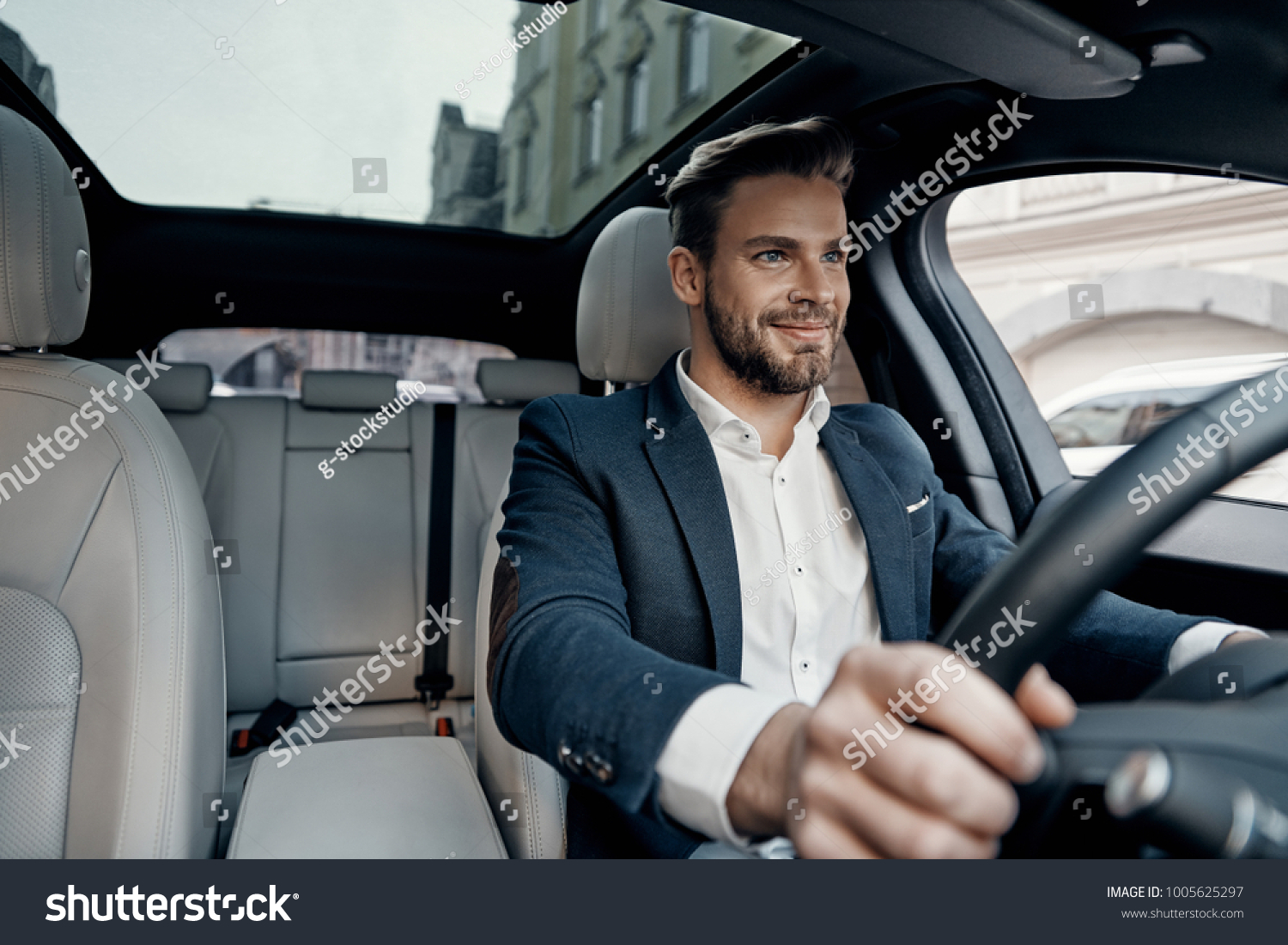 Success in motion. Handsome young man in full suit smiling while driving a car #1005625297