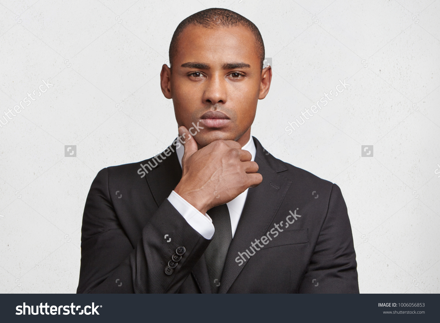 Indoor shot of serious mixed race male magnat dressed in elegant black suit, looks confidently into camera, waits for business partner as going to sign indemnity contract, isolated over white wall #1006056853