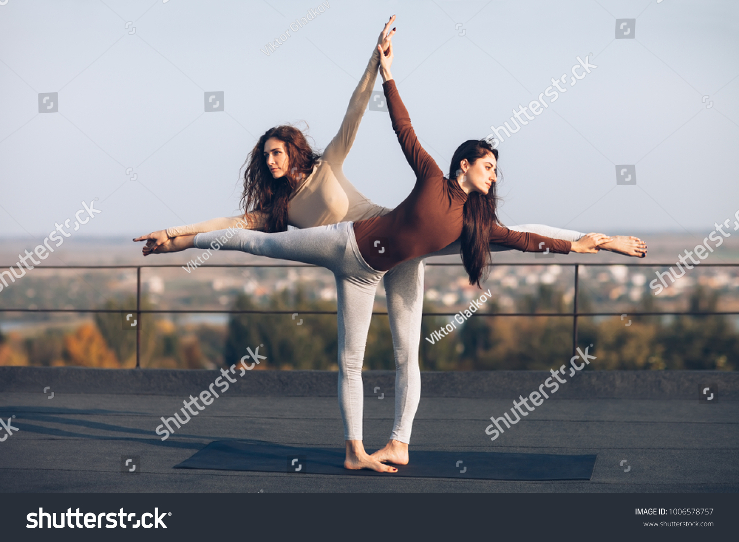 Two young beautiful women doing yoga asana virabhadrasana helping each other on the roof outdoor. partner yoga. Balance, concentration, equilibrium concept #1006578757