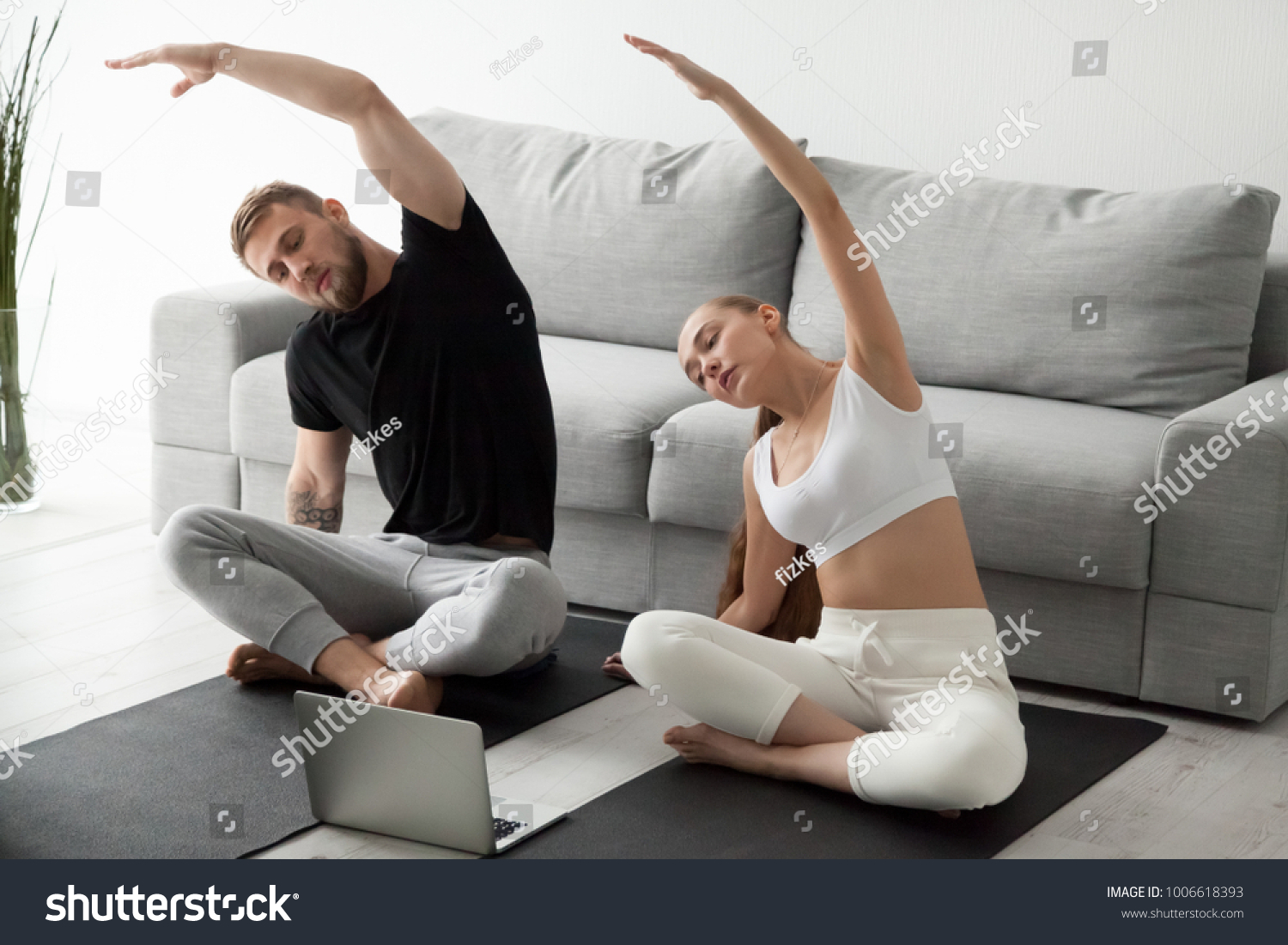 Sporty young couple warming up stretching watching fitness video tutorial online on laptop, fit man and woman doing workout at home sitting on mat in living room practicing yoga side bending exercise #1006618393