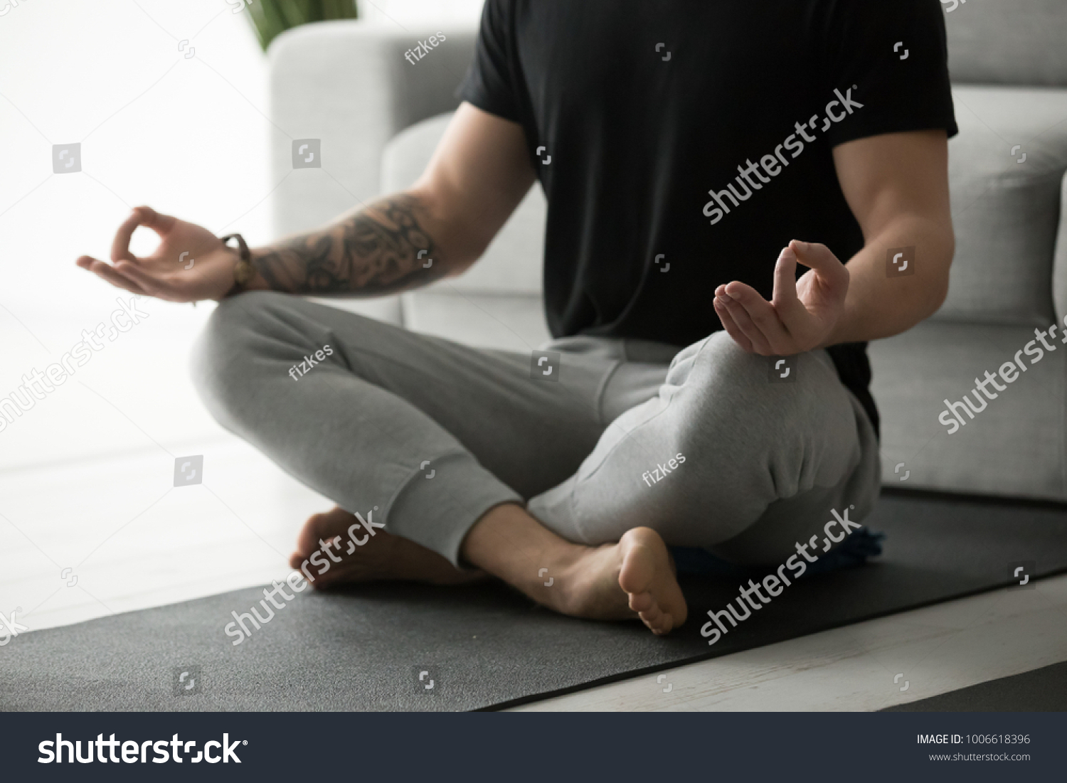 Sporty mindful man with tattoo meditating alone at home, peaceful calm hipster fit guy practicing yoga in lotus pose indoors holding hands in mudra, freedom and calmness concept, close up view #1006618396