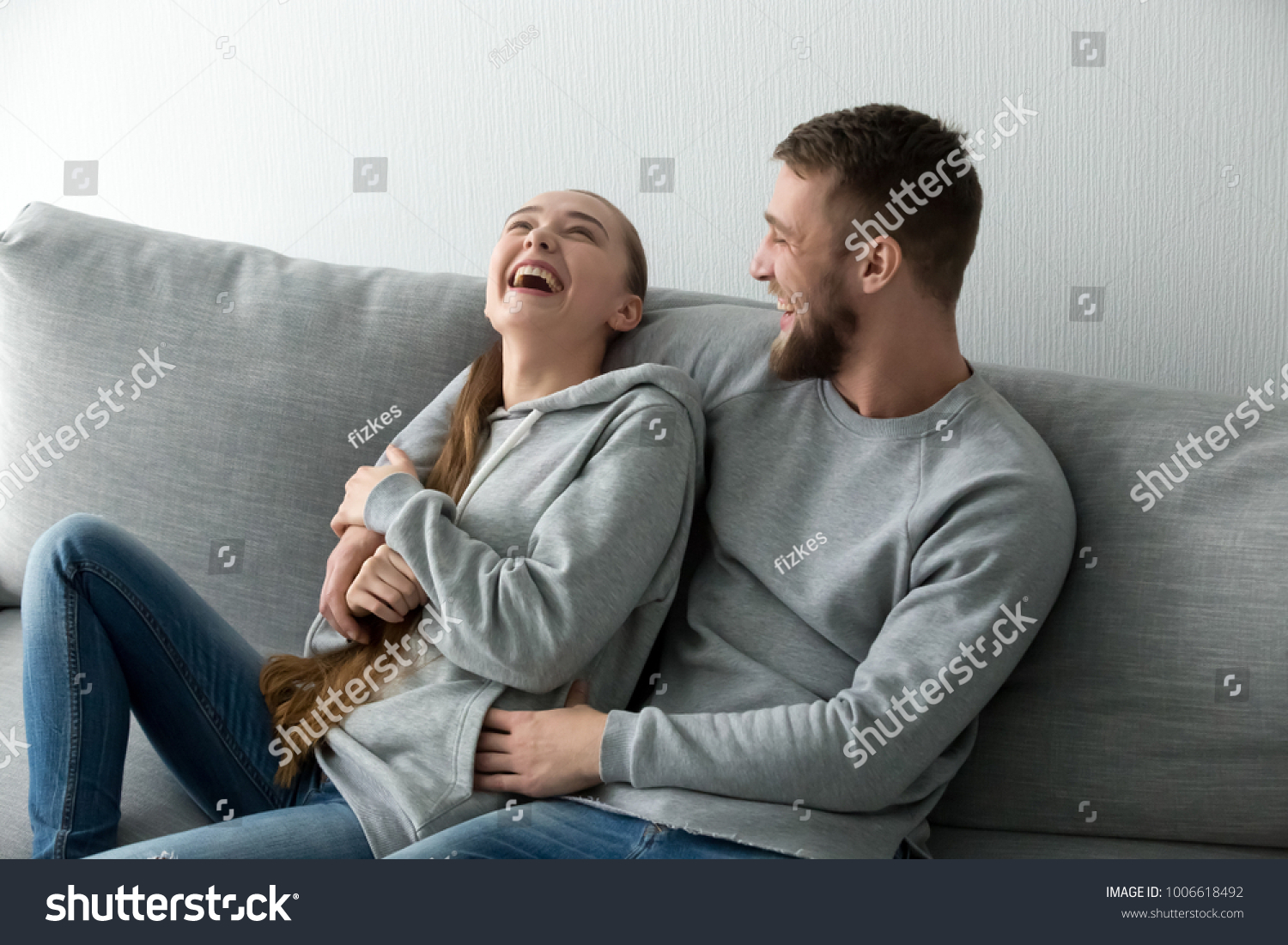 Young happy couple having fun talking laughing relaxing at home on couch, boyfriend embracing girlfriend telling funny joke sitting on sofa, humor in relationships, enjoying weekend together #1006618492