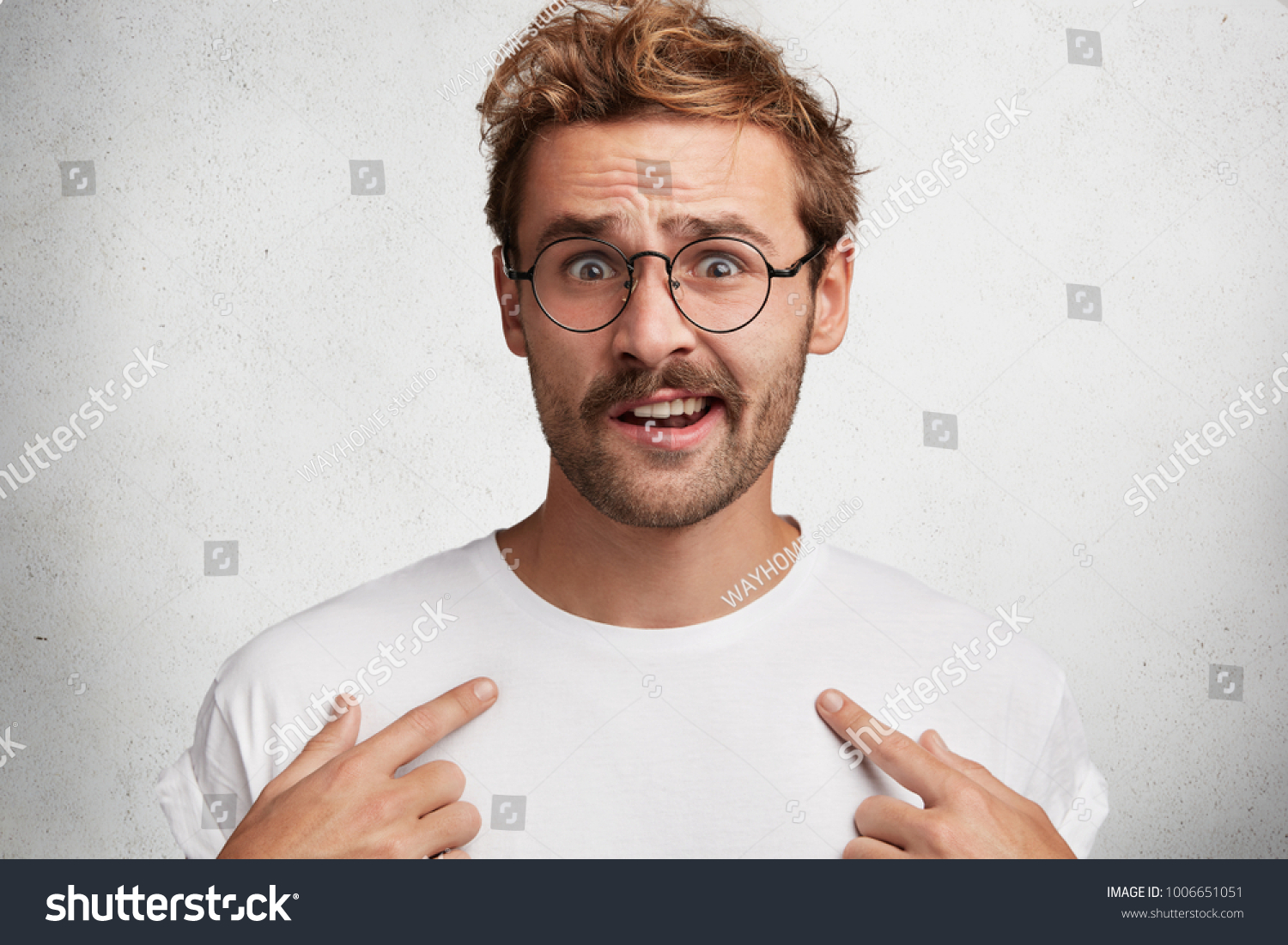 Isolated shot of indignant male with trendy hairstyle indicates at blank copy space of white t shirt, can`t understand something, advertises clothes. People, design, clothing and fashion concept #1006651051