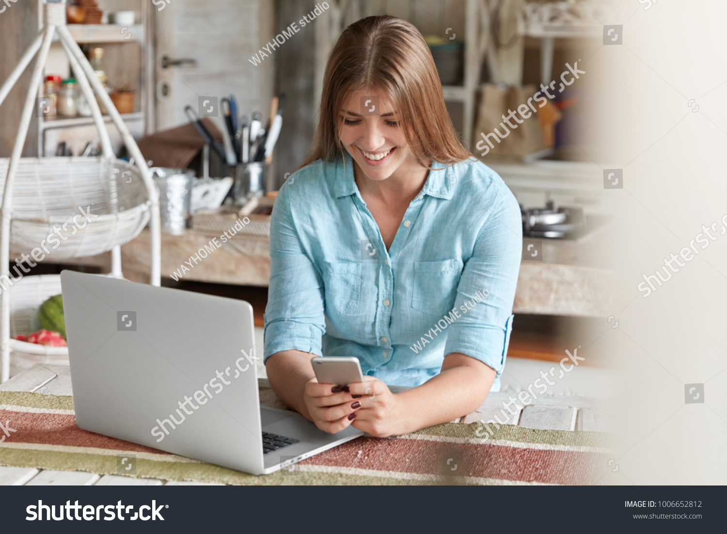 Satisfied young female freelancer works remotely, sits against kitchen interior, glad to recieve message on smart phone, types answer. Modern technology, communication and lifestyle concept. #1006652812