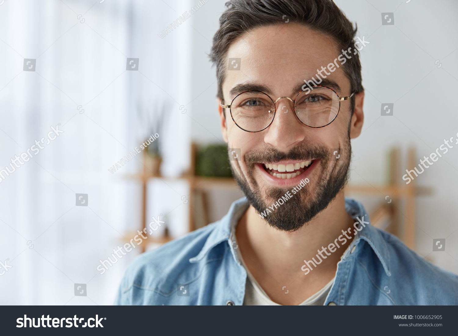 Close up shot of cheerful satisfied attractive male with stubble, has broad smile, wears round spectacles, rejoices success at work, stands against cozy interior. Fashionable designer glad be praised #1006652905
