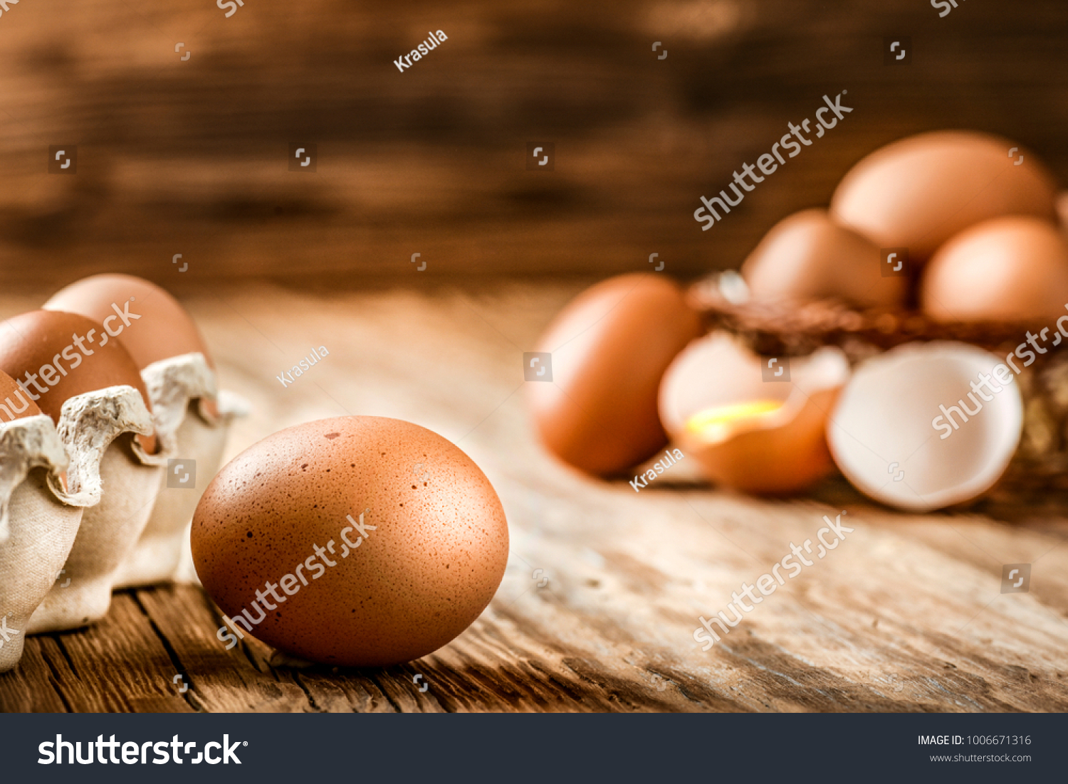 Brown eggs in carton box. Broken egg with yolk in background.  #1006671316