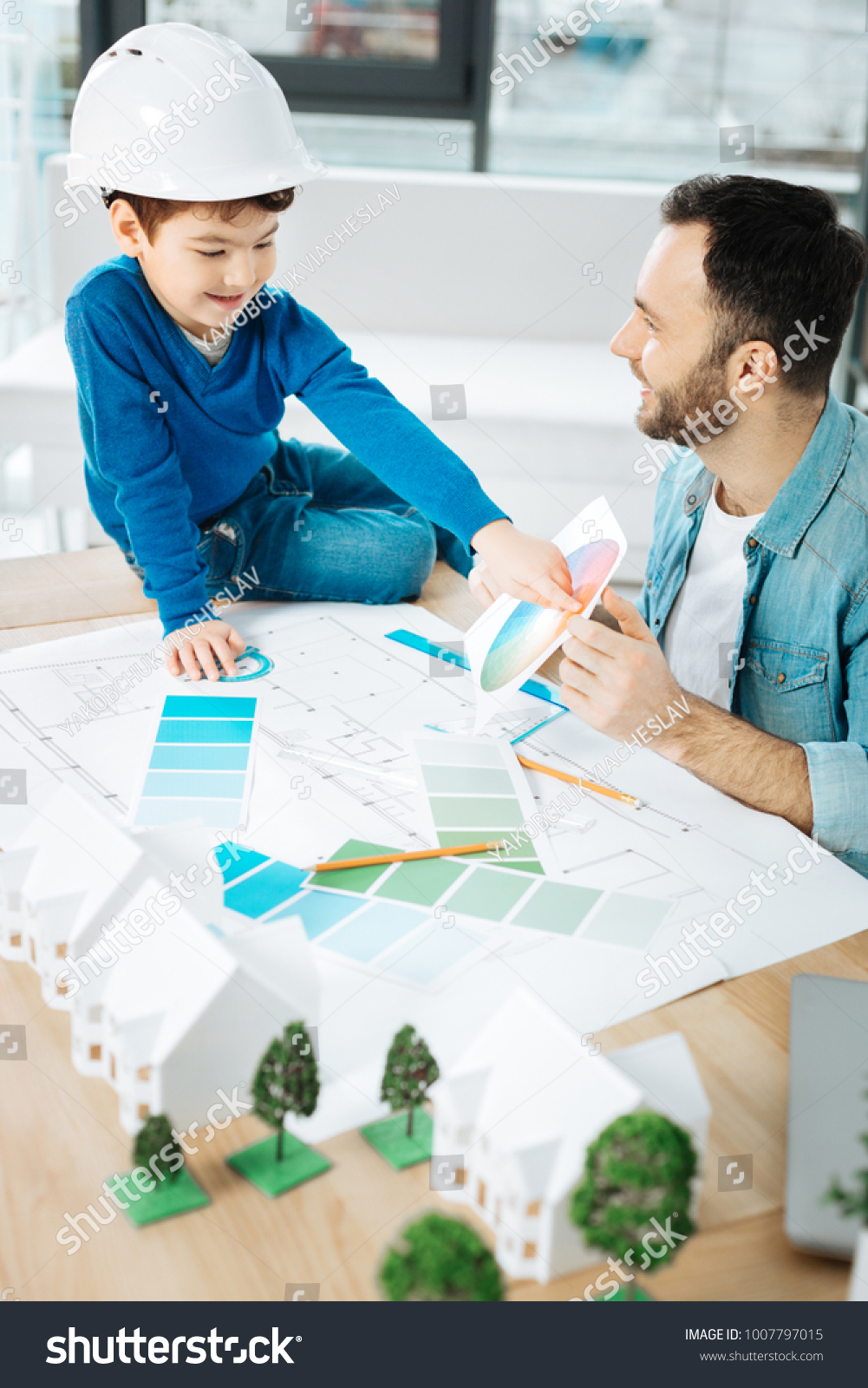 Crucial advice. Charming young architect sitting next to his little son and discussing the color choice with him while the boy pointing at color in the color chart #1007797015