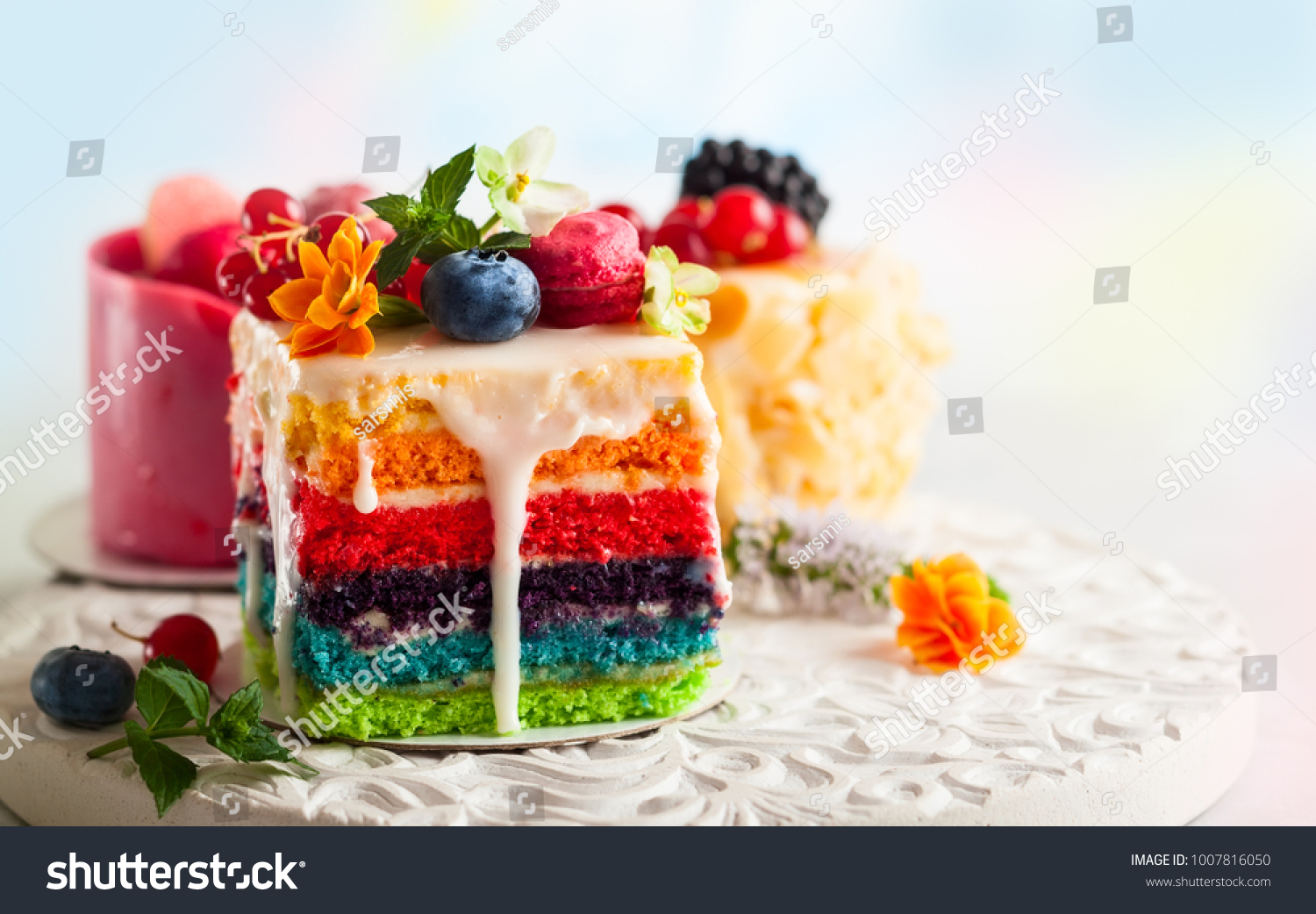 Various slices of cakes on a white tray: rainbow cake, raspberry cake and almond cake. Sweets decorated with fresh berries and flowers for holiday #1007816050