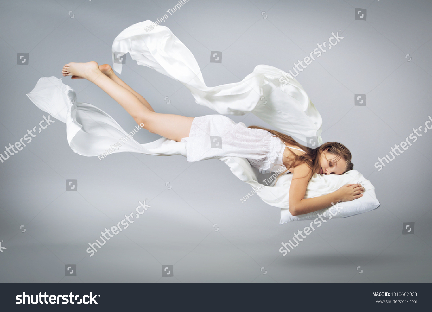 Sleeping girl. Flying in a dream. White linen flying through the air. Light grey background #1010662003