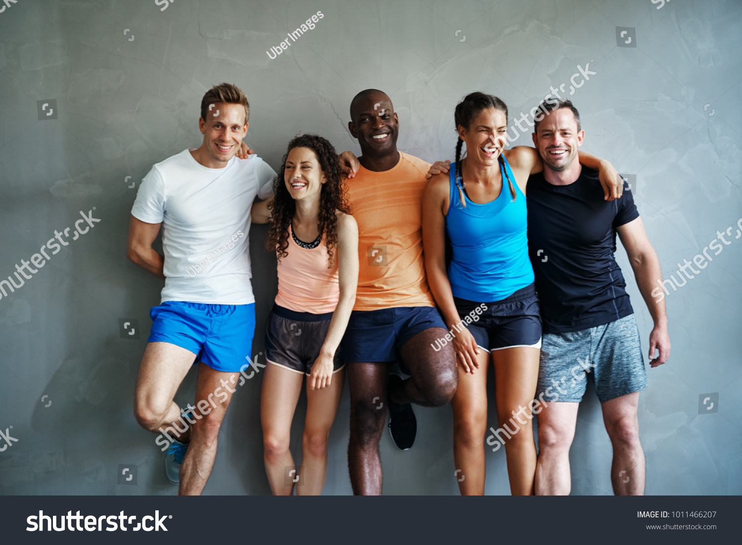 Smiling group of sporty friends in sportswear laughing while standing arm in arm together against the wall of a gym after a workout #1011466207