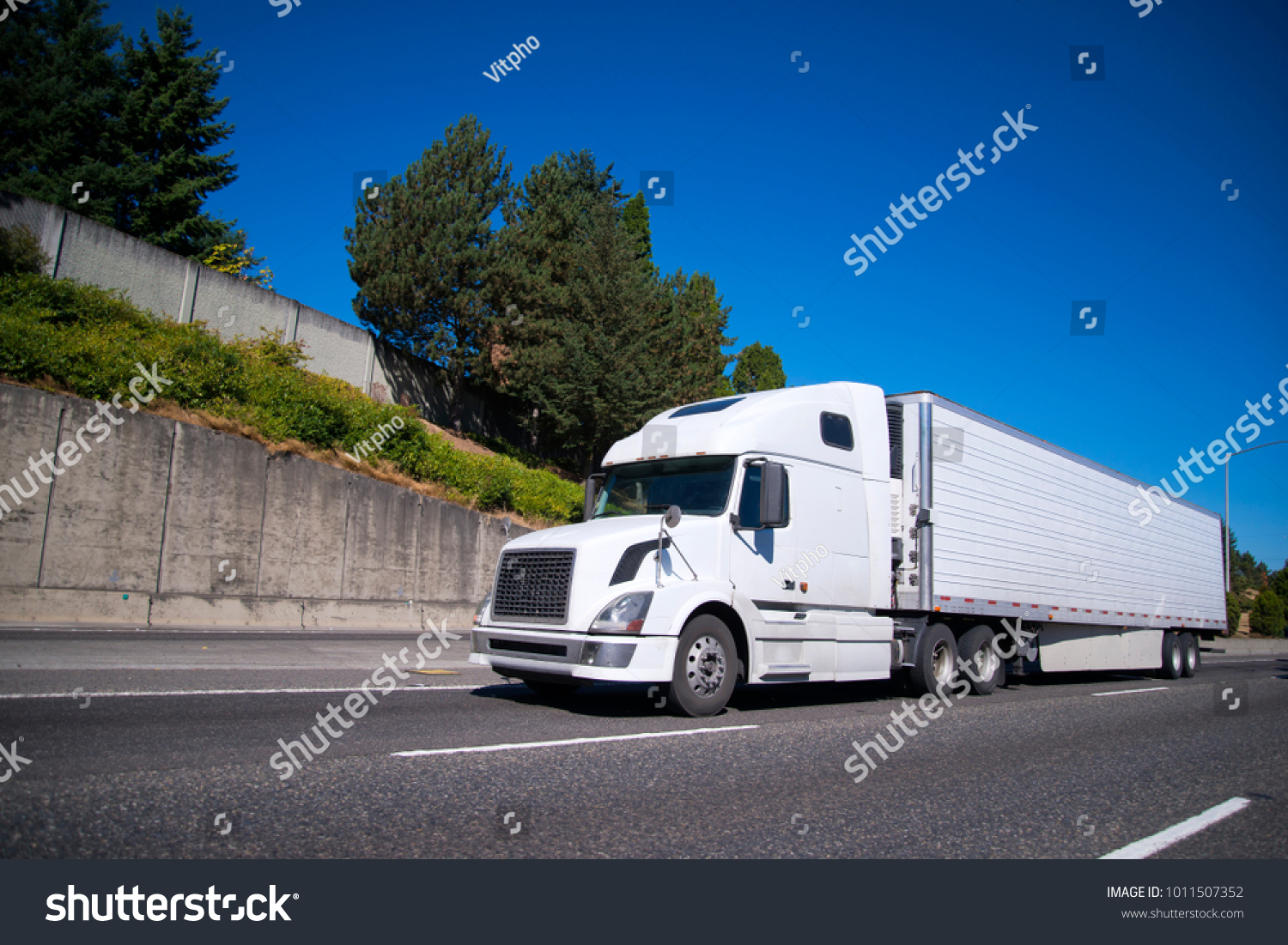 White modern Big rig semi truck with reefer semi trailer equipped with refrigeration unit going on flat wide highway with commercial cargo for delivery to warehouse #1011507352