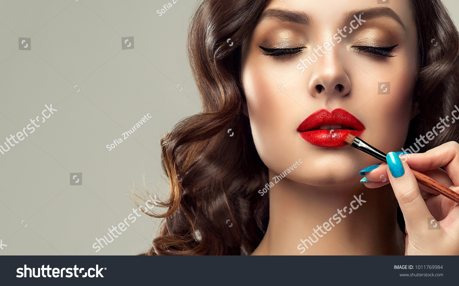  Makeup artist applies  red lipstick  . Beautiful woman face. Hand of make-up master, painting lips of young beauty  model girl . Make up in process #1011769984