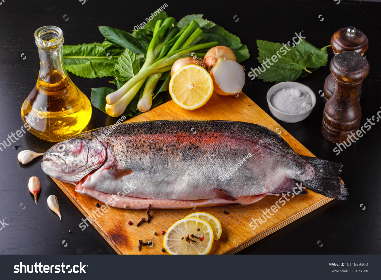Fresh gutted trout on a wooden cutting board surrounded by ingredients for baking: olive oil, lemon, spring onions, garlic and spices. Top view #1011829303