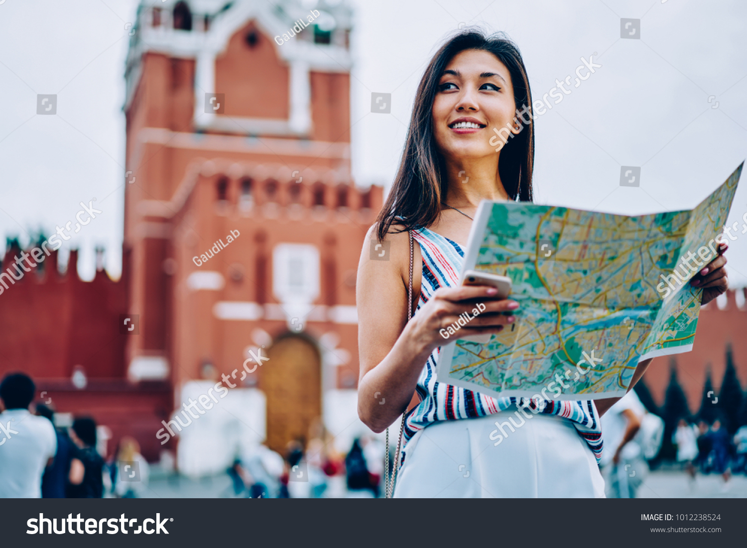 Attractive asian female traveler standing on red square in Moscow enjoying sightseeing tour with map,cheerful woman tourist getting to location near Kremlin on background planning next route in Moscow #1012238524