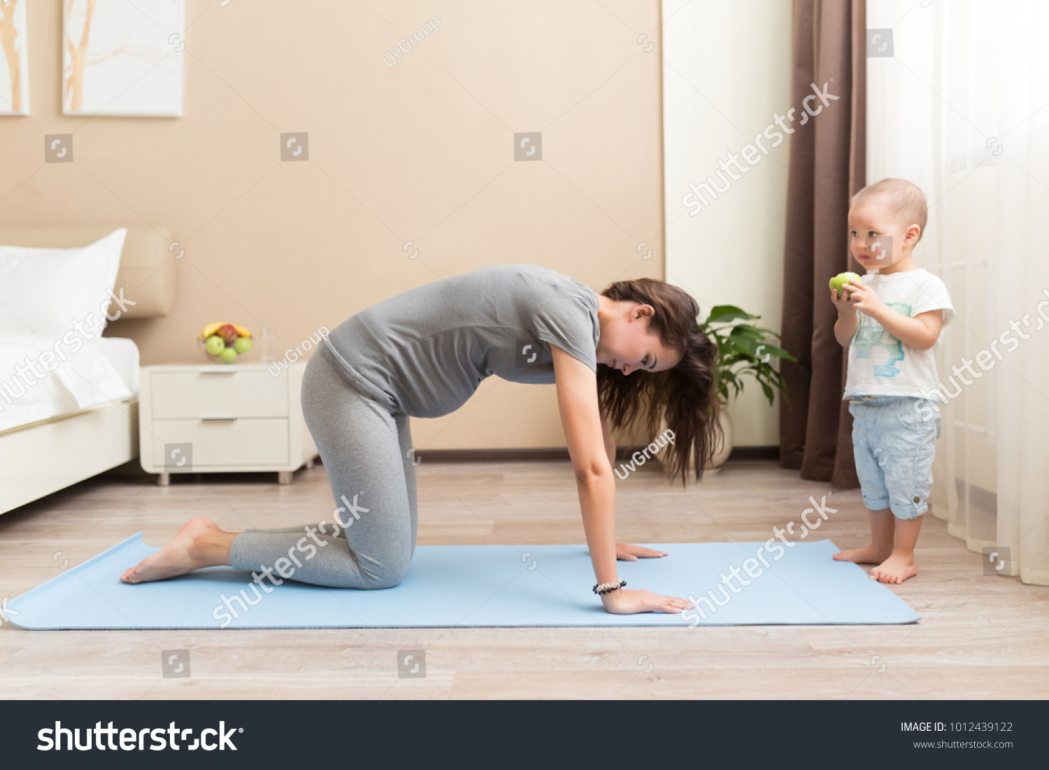 Beautiful young pregnant woman and little kid boy smiling while lying on yoga mat and doing fitness exercises at home #1012439122
