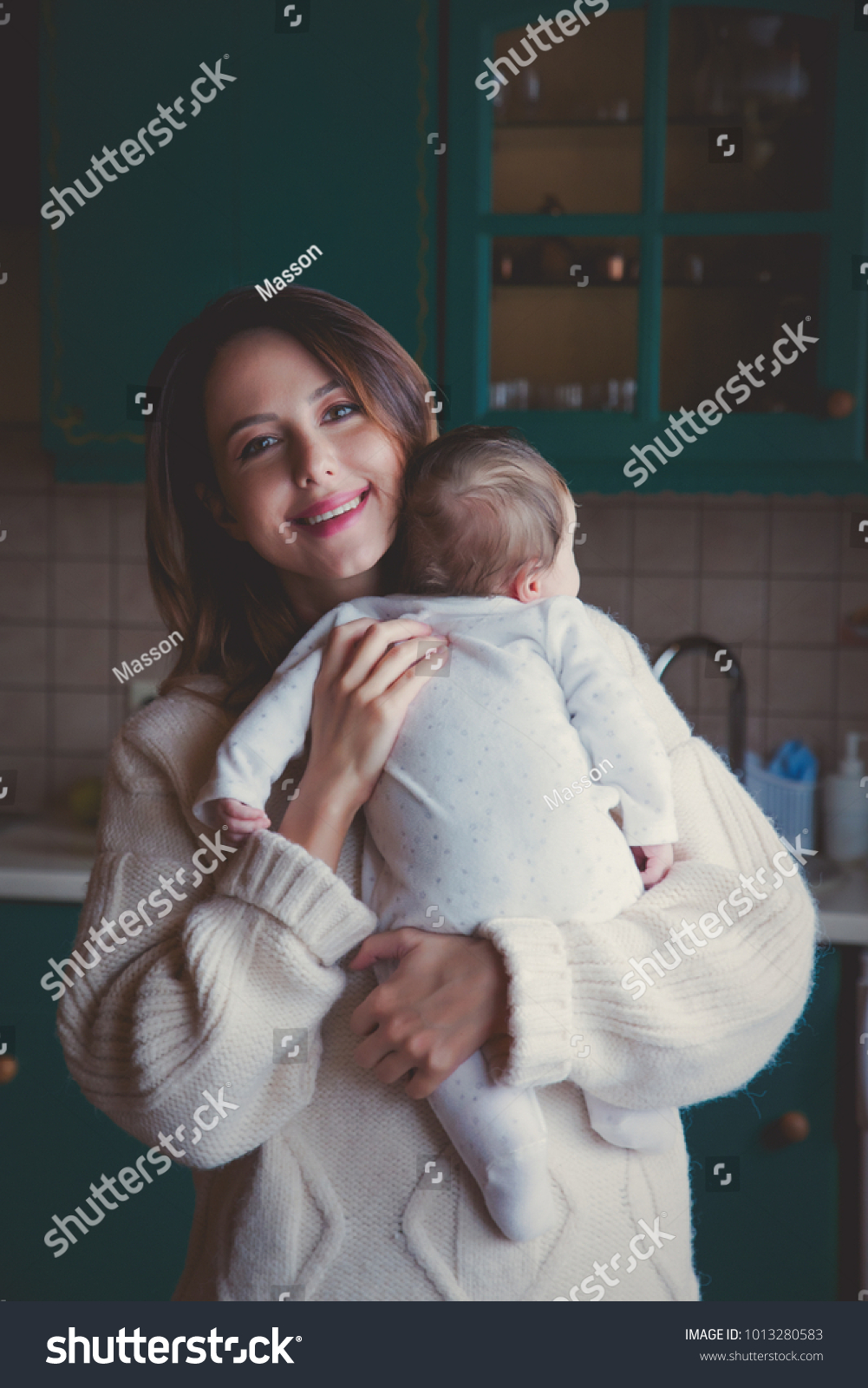 Young mother in sweater holding child indoor at kitchen.  #1013280583