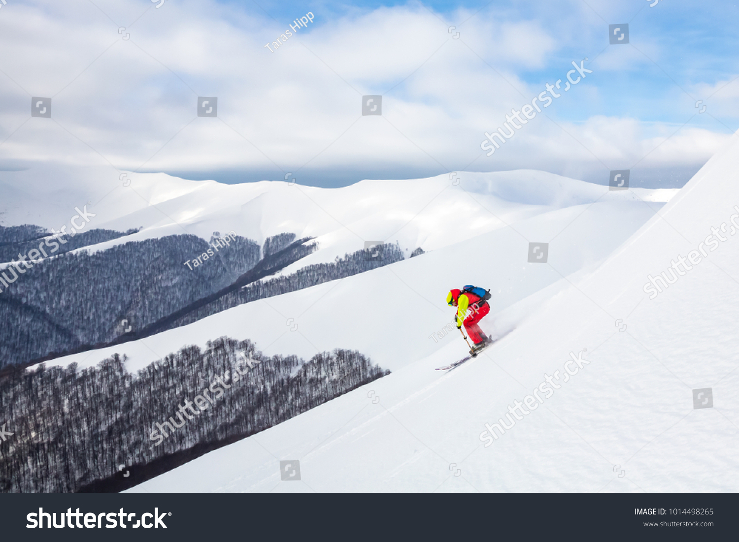 A man is skiing on the slope, wearing brigh  red yellow jumpsuit. Good skiing day, blue sky on the background. #1014498265