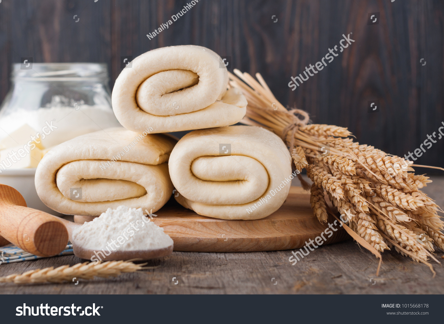 Puff pastry dough. Homemade folded raw puff pastry on a wooden cutting board on a rustic wooden surface (table). Making puff pastry. Dough's rolls with a rolling pin, flour, butter, wheat spikelets. #1015668178