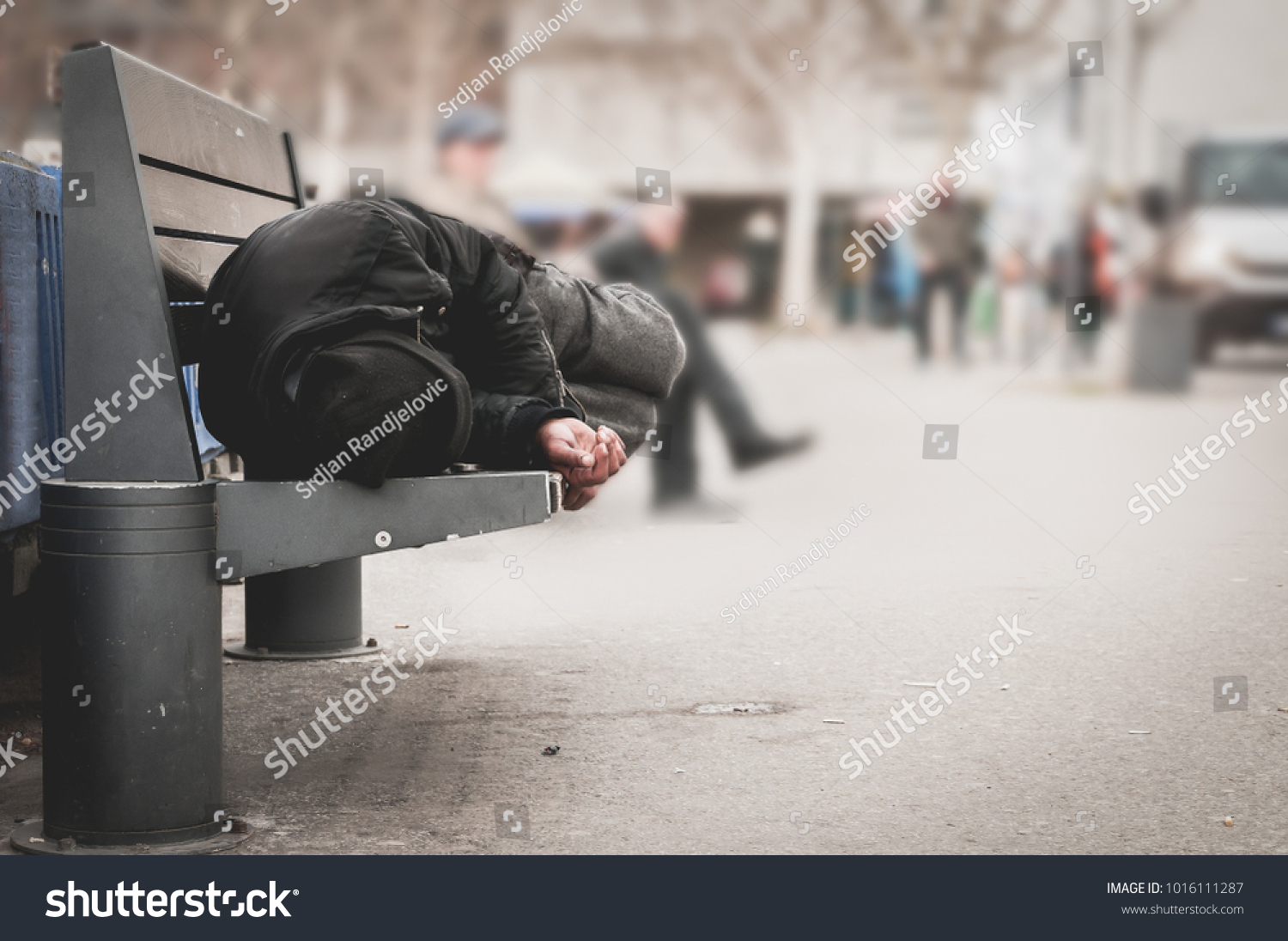 Poor tired depressed hungry homeless man or refugee sleeping on the wooden bench on the urban street in the city, social documentary concept #1016111287