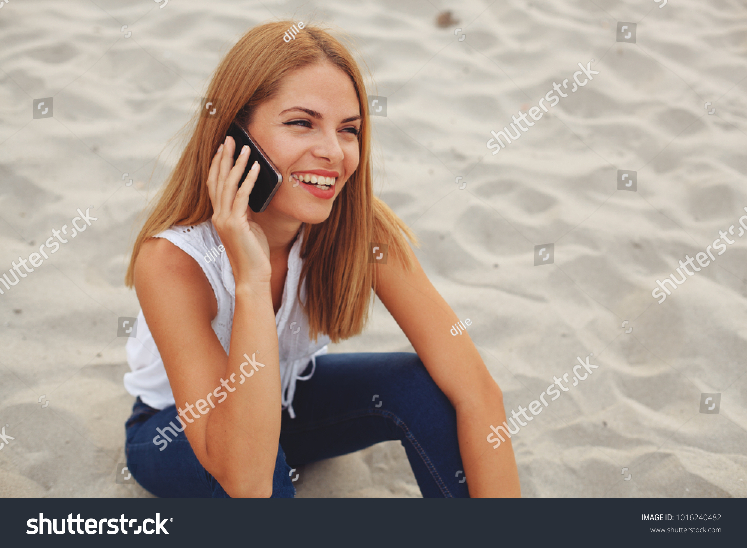 Young happy woman sitting on the beach sand and talking on the phone #1016240482