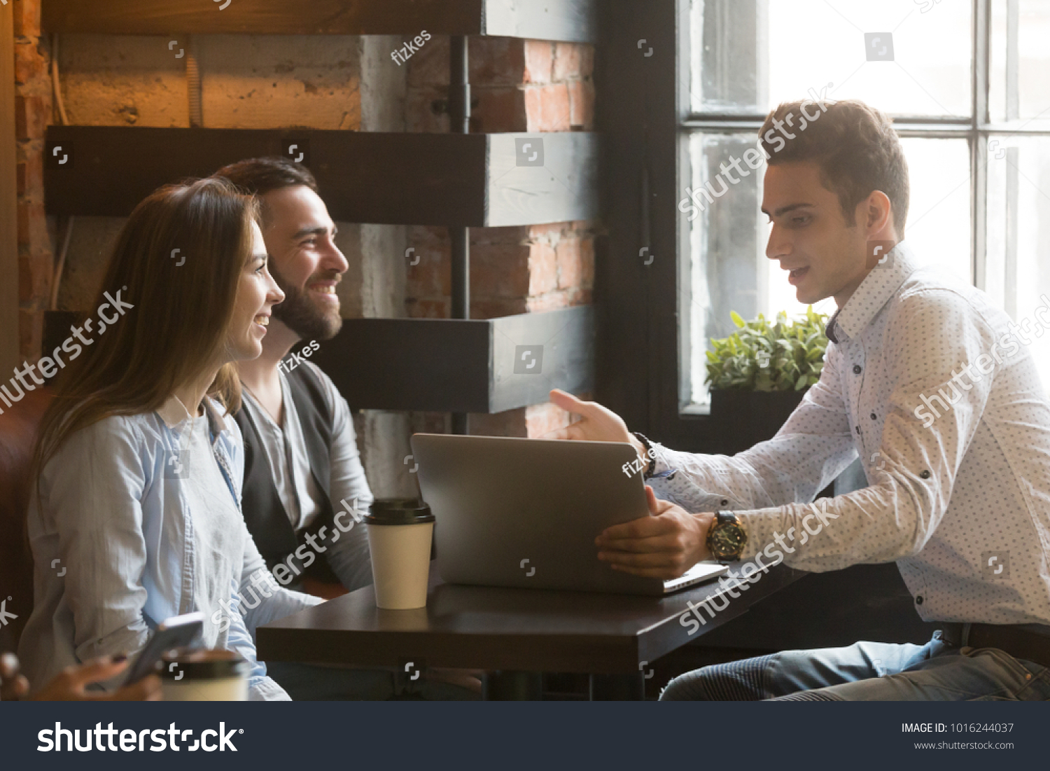 Insurance broker or salesman making offer to young millennial couple using laptop in cafe, realtor consulting customers about mortgage sitting at coffeehouse table pointing on computer screen #1016244037