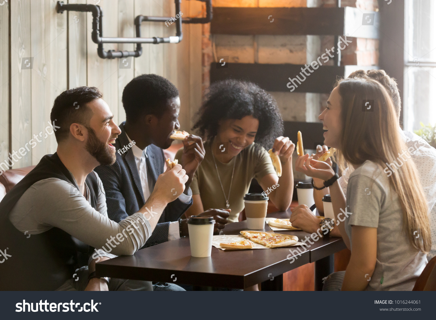 Multiracial happy young people eating pizza in pizzeria, black and white cheerful mates laughing enjoying meal having fun sitting together at restaurant table, diverse friends share lunch at meeting #1016244061