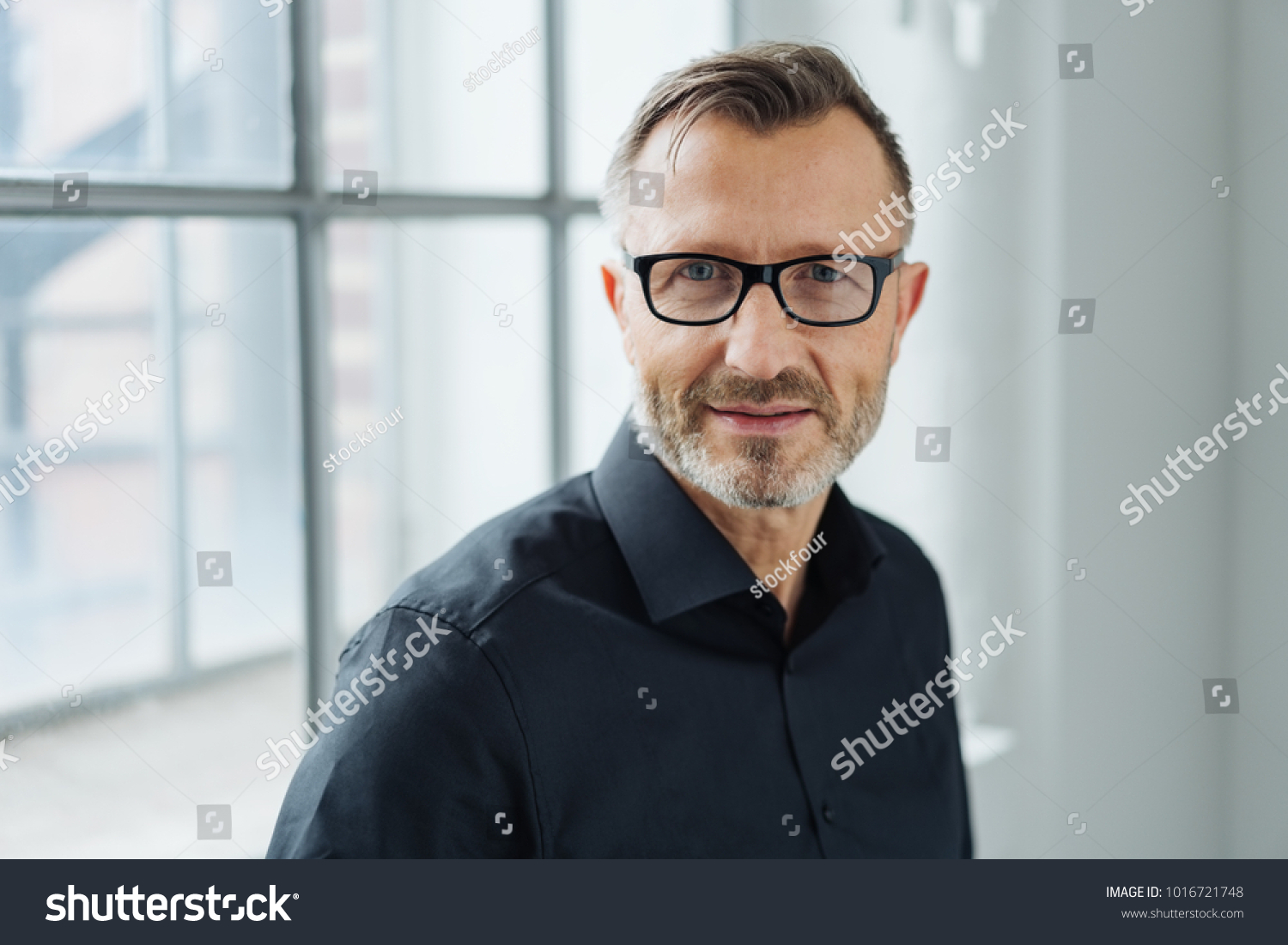 Close-up portrait of a middle-aged man wearing black shirt and eyeglasses while looking at camera with confidence in the office #1016721748