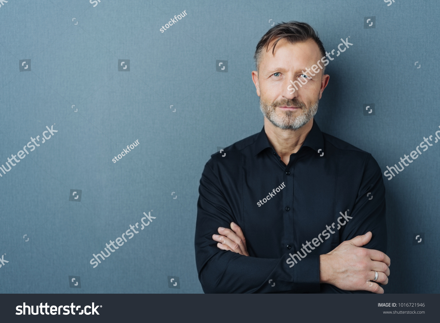 Serious middle-aged man with folded arms and a deadpan expression posing in front of a grey background with copy space #1016721946